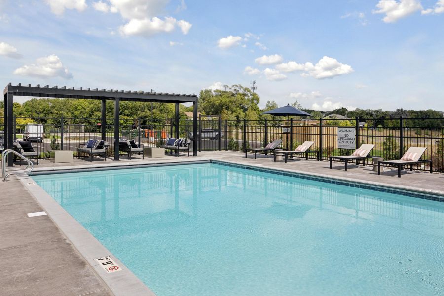 Outdoor swimming pool with lounge chairs, pergola shade, and a black fence on a sunny day.