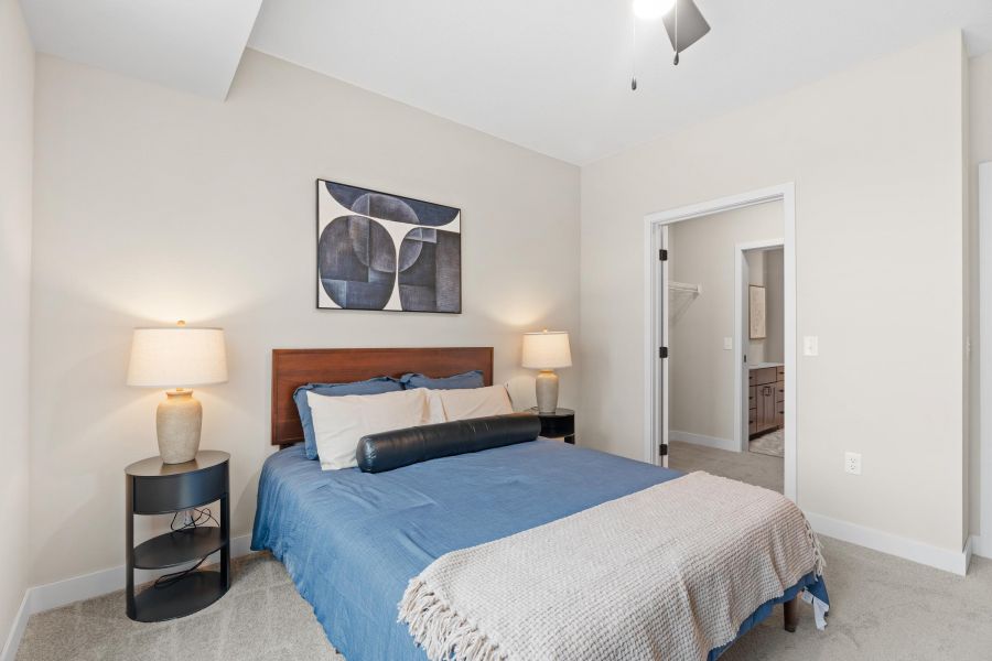 Modern bedroom with blue bedding, two bedside lamps, and abstract art above the bed. Closet and bathroom visible.