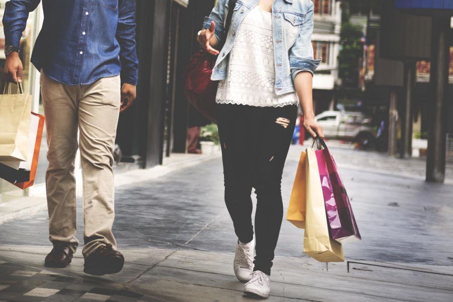 Two people walking outdoors, carrying multiple shopping bags, with storefronts in the background.