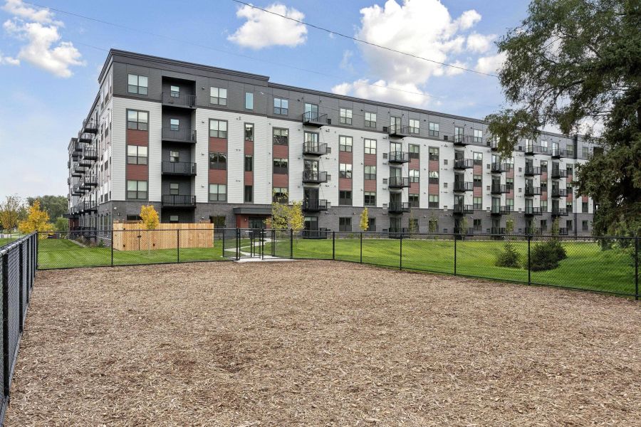 A fenced dog park with wood chips in front of a modern apartment building on a clear day.