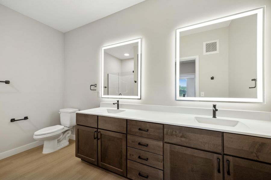 Modern bathroom with dual sinks, lighted mirrors, wooden cabinets, and a toilet on a light wood floor.