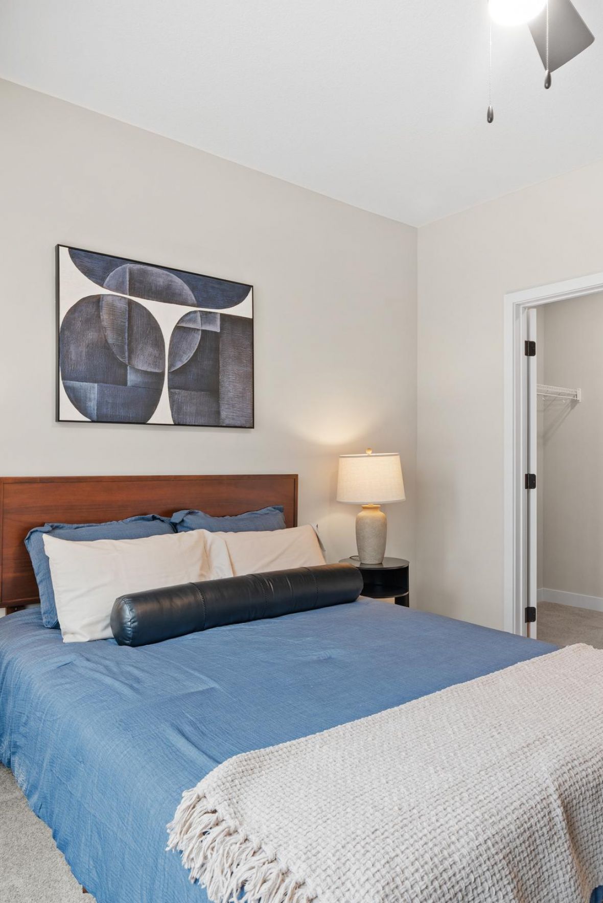 Modern bedroom with blue bedding, two bedside lamps, and abstract art above the bed. Closet and bathroom visible.