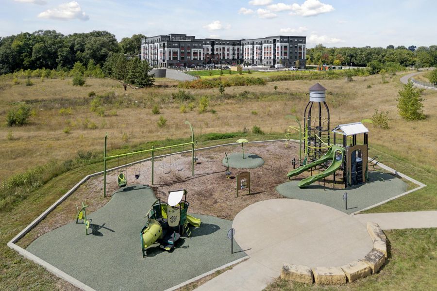 A playground with green equipment sits in an open field, with an apartment building in the background.