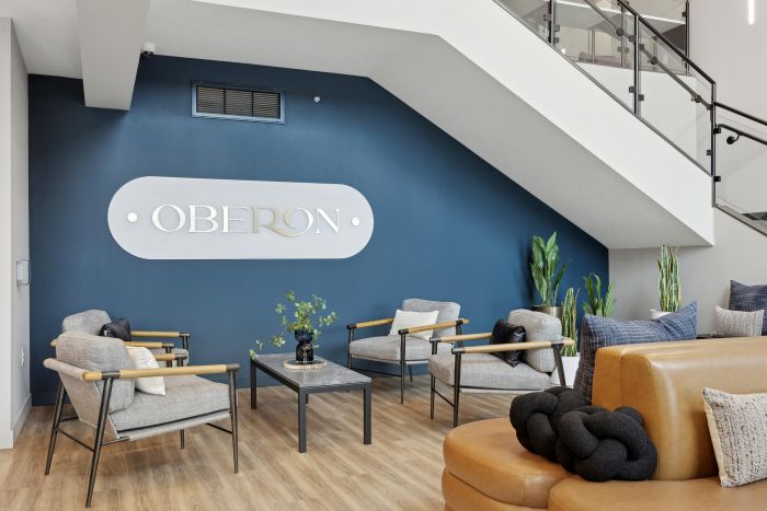 Modern lobby with gray chairs, a tan sofa, plants, and "Oberon" sign on a navy blue wall under stairs.