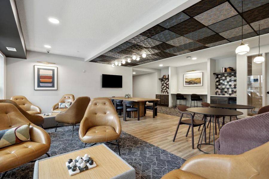 Modern lounge with tan chairs, tables, abstract art, and a TV on the wall in a bright, open space.