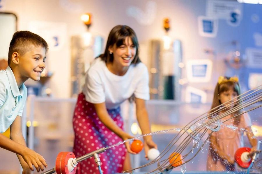 Two children and a woman play with a water-based interactive exhibit indoors, smiling and having fun.