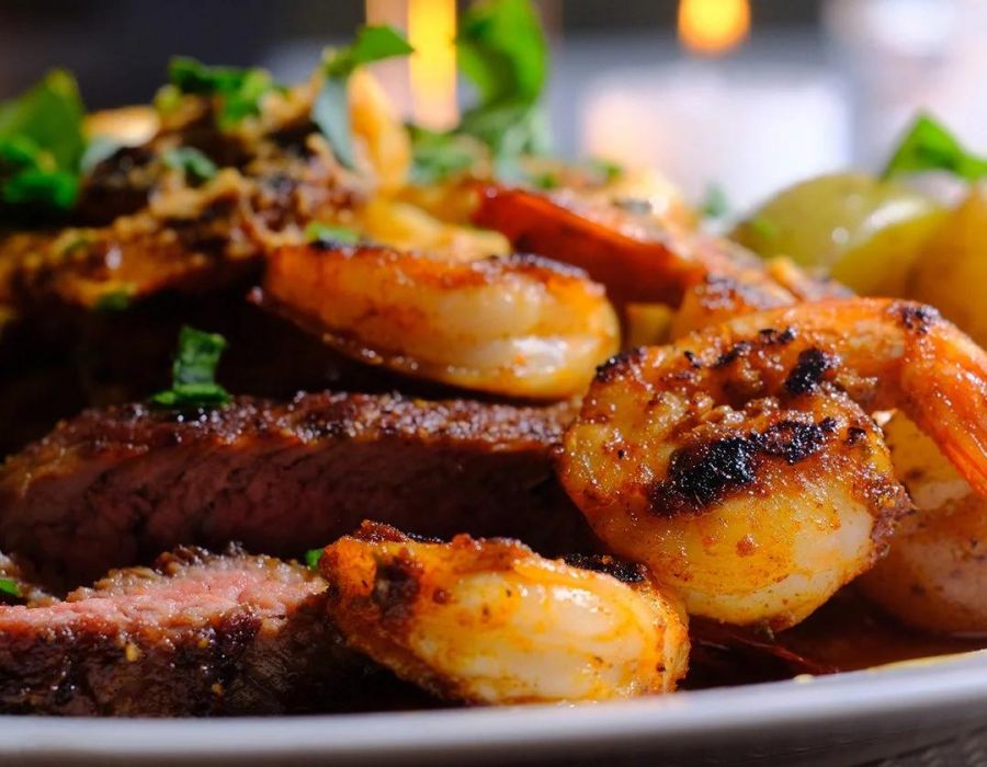 Grilled shrimp and steak served with potatoes and garnished with herbs on a white plate.