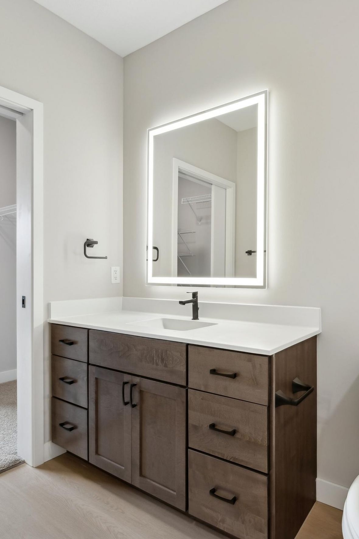 Modern bathroom vanity with brown cabinets, a white countertop, and a large backlit mirror above the sink.