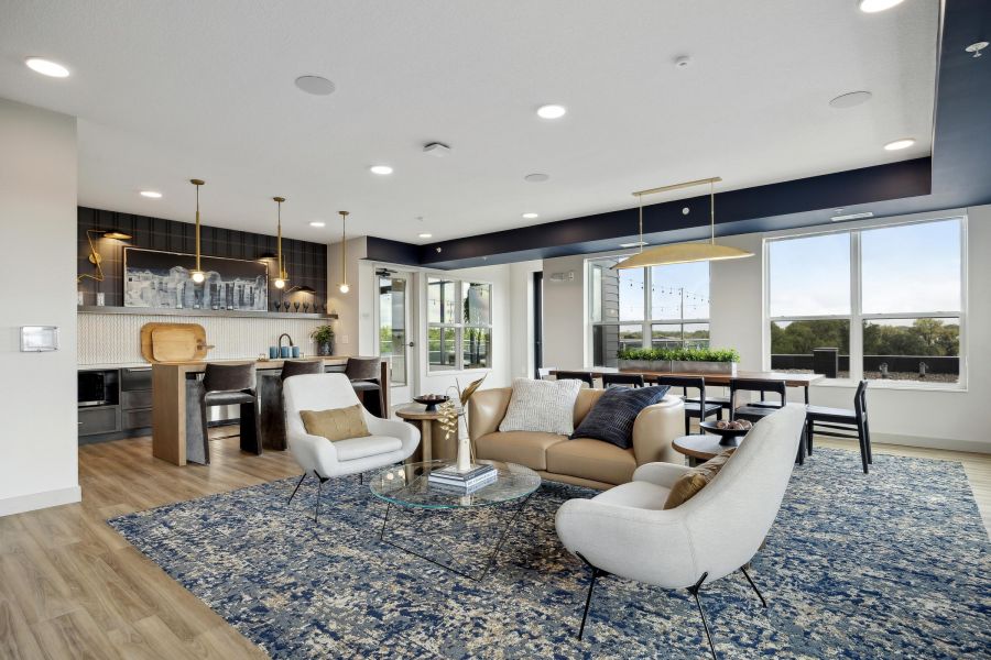 Modern living room with a blue rug, light furniture, large windows, and an open kitchen in the background.