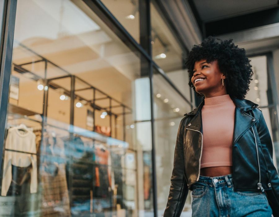 Smiling woman in a leather jacket walking past a clothing store with glass windows.