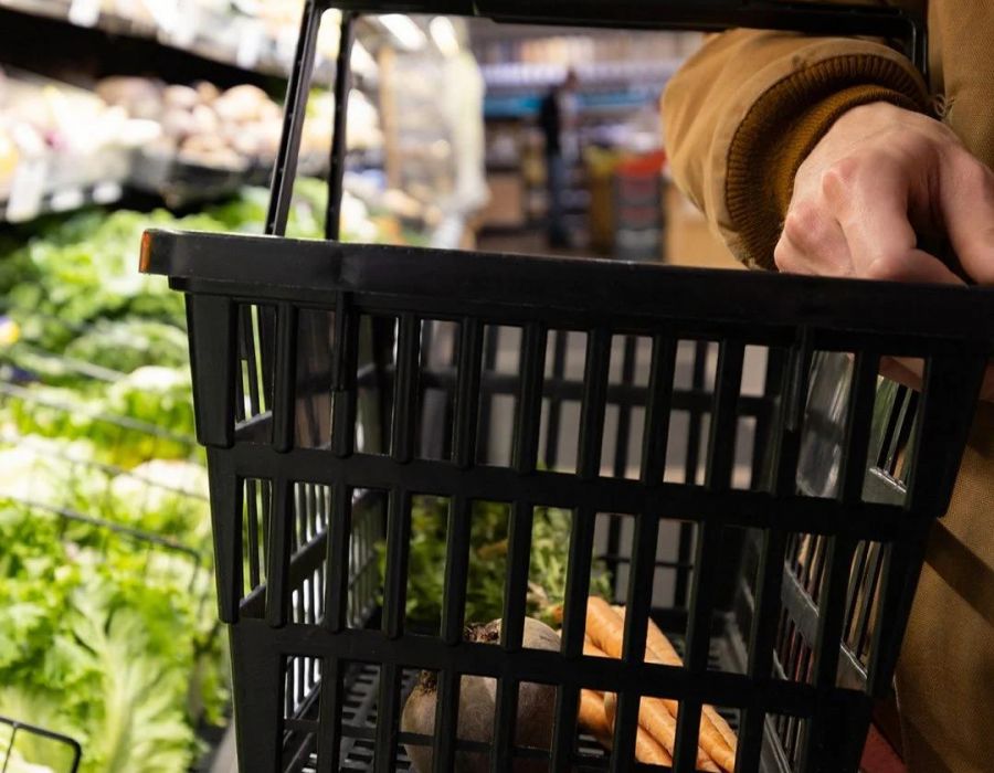A person holding a shopping basket with vegetables in a grocery store produce aisle.
