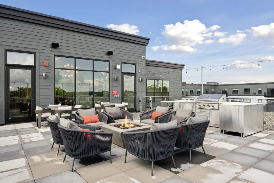 Modern rooftop patio with a fire pit, lounge chairs, barbecue grill, and string lights under a blue sky.