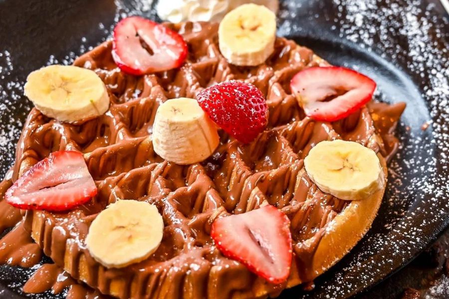 Waffle topped with chocolate, banana slices, strawberries, whipped cream, and powdered sugar on a black plate.