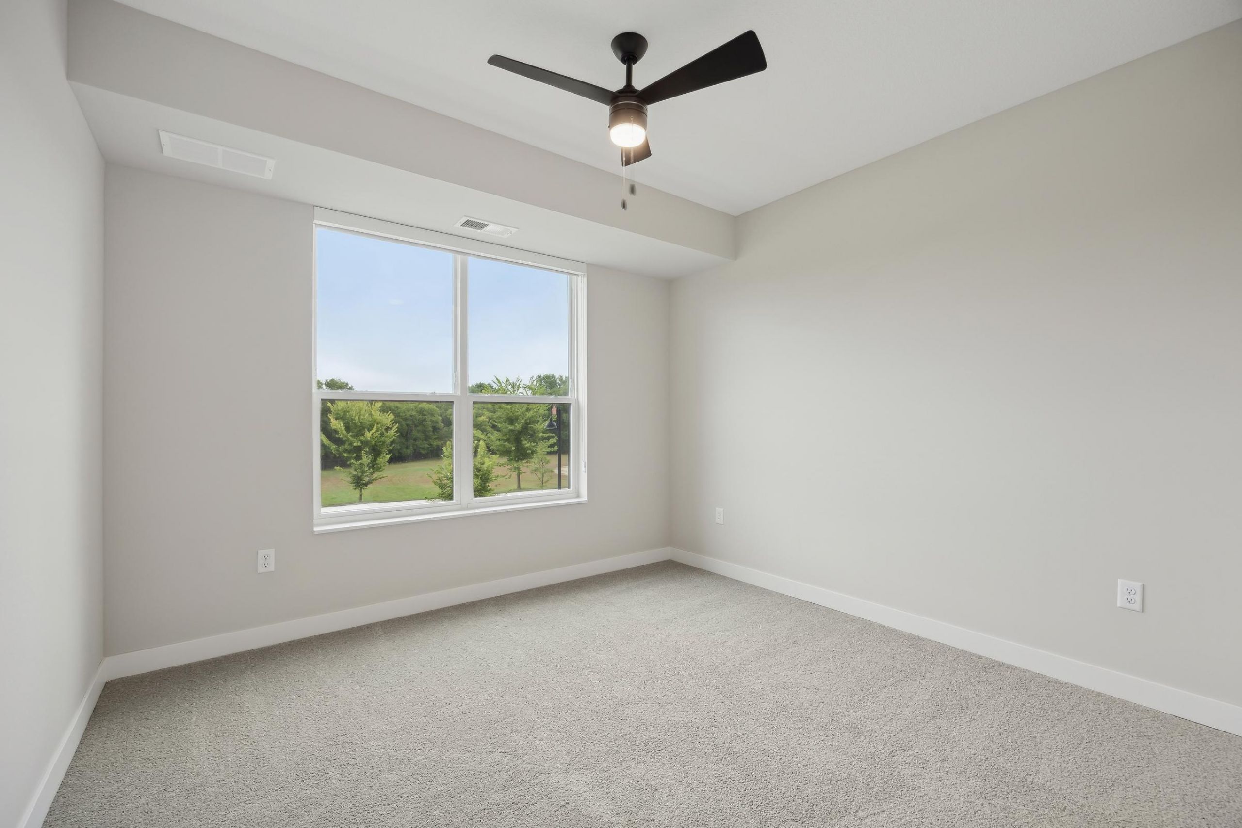 Empty bedroom with beige carpet, large window, ceiling fan, and light gray walls.