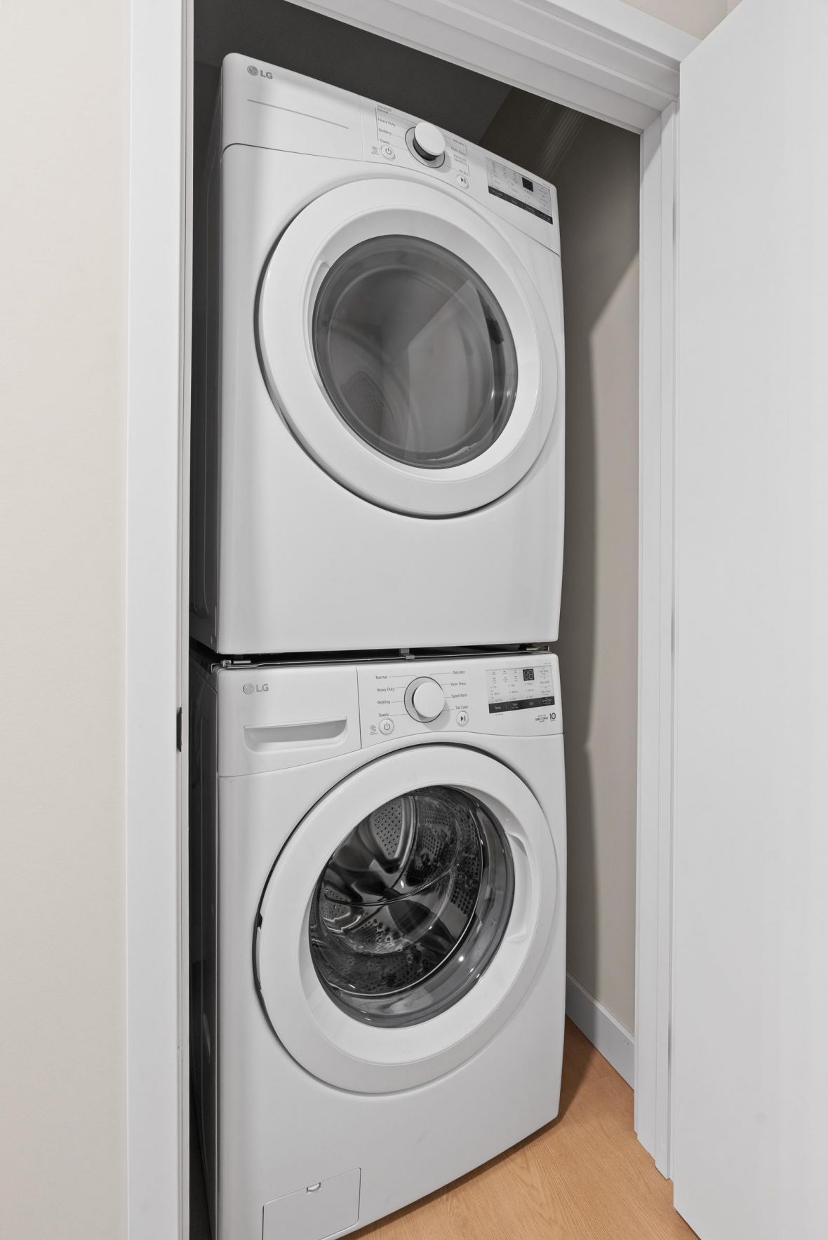 Stacked white washer and dryer set inside a small, white-walled laundry closet with a light wood floor.