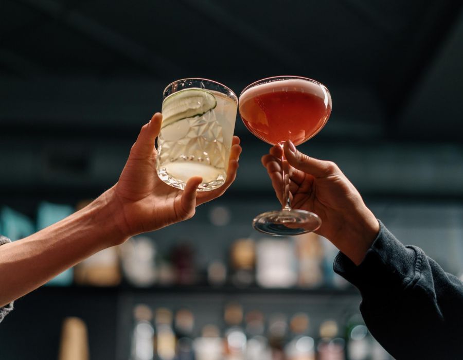 Two people clinking cocktail glasses in a toast, with a blurred bar background.