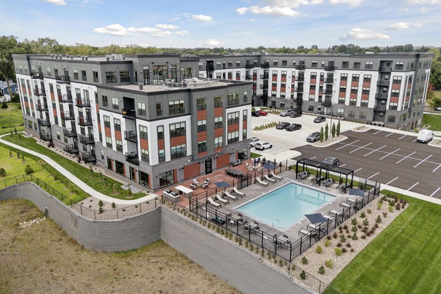 A modern apartment complex with a pool, sun loungers, parking lot, and landscaped green areas.