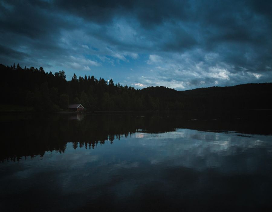 A small cabin sits by a calm lake, surrounded by trees and under a cloudy, darkening sky at dusk.