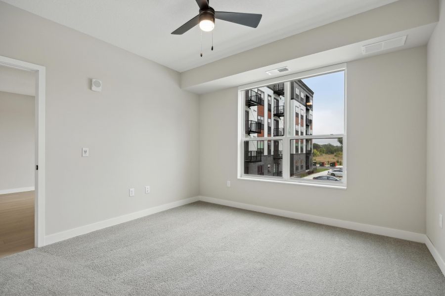 Empty, carpeted bedroom with large window, ceiling fan, and view of a modern apartment building outside.