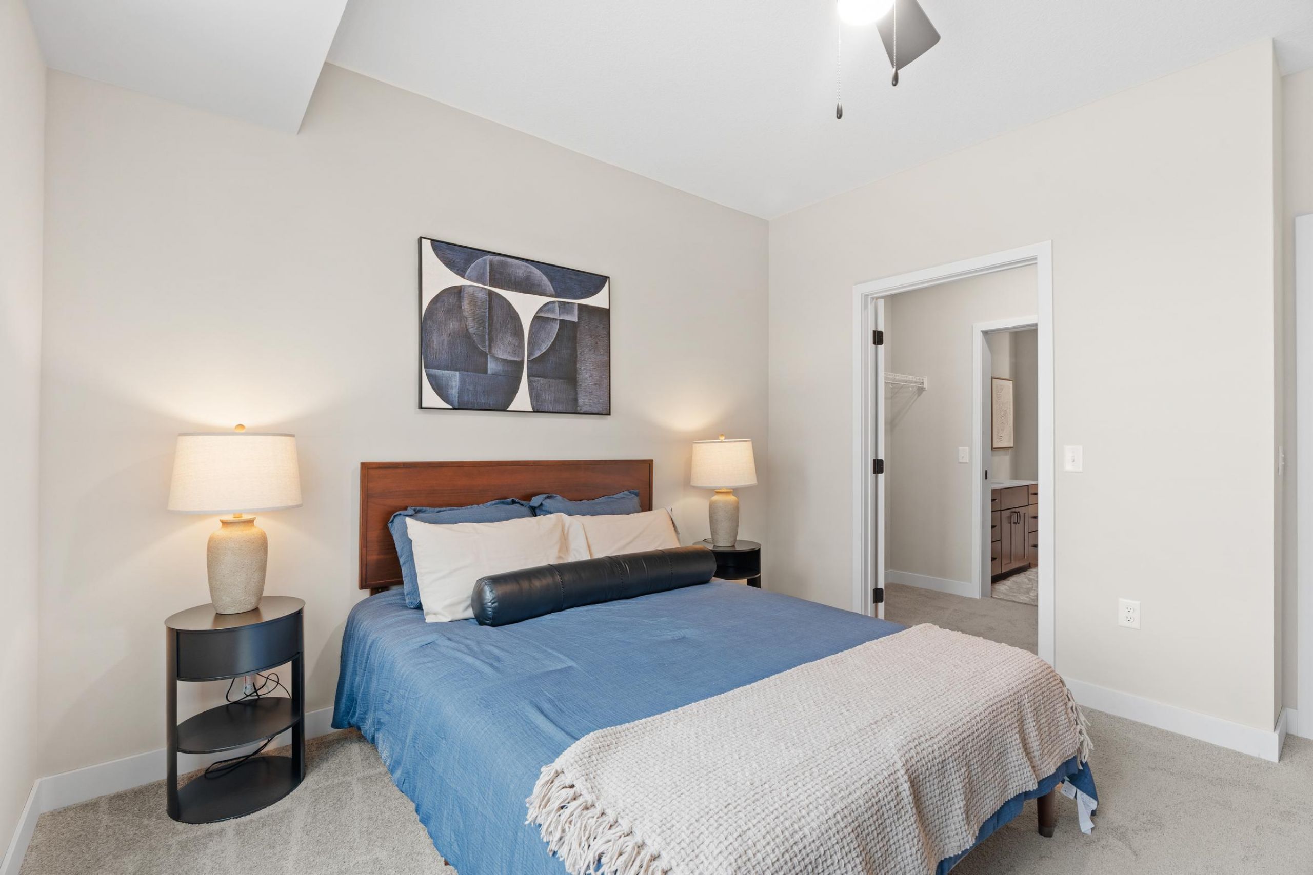 Modern bedroom with blue bedding, two bedside lamps, and abstract art above the bed. Closet and bathroom visible.