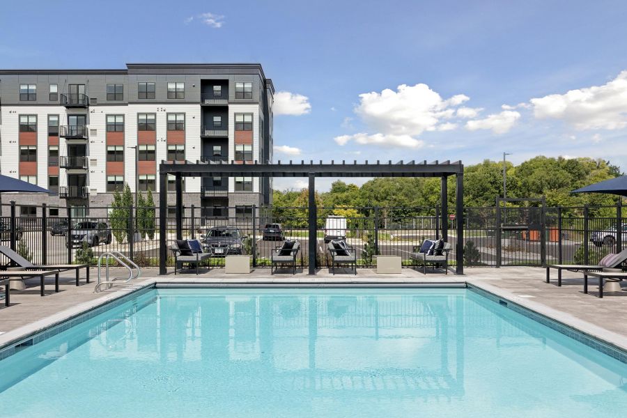 Swimming pool with lounge chairs and pergolas, set in front of a modern apartment building on a sunny day.