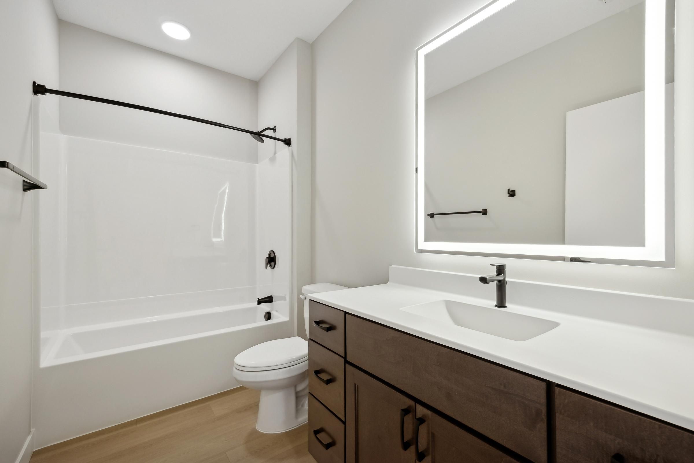 Modern bathroom with a white tub, toilet, dark wood vanity, and illuminated mirror.