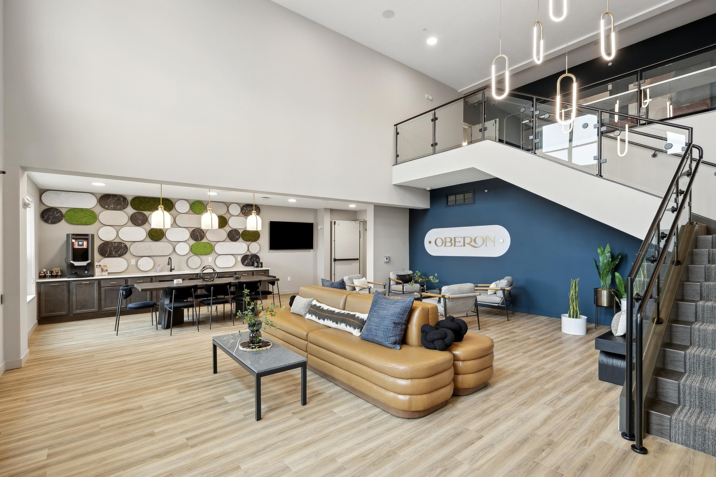 Modern lobby with tan sofas, a meeting table, plants, and a staircase leading to an upper floor.