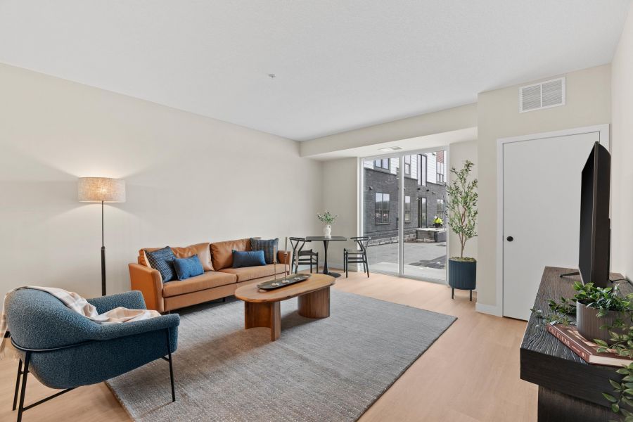 Modern living room with a tan sofa, blue chair, coffee table, TV, and large window letting in natural light.