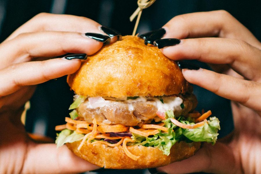Hands with black nail polish holding a sandwich with lettuce, carrot, and a round bun on a skewer.