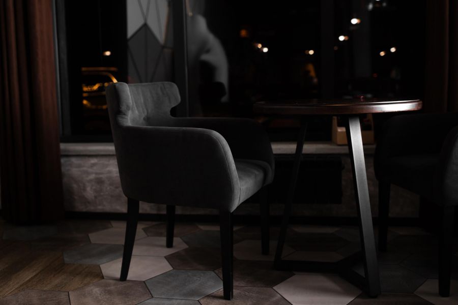 A dark, modern chair sits next to a round wooden table in a dimly lit room with geometric floor tiles.