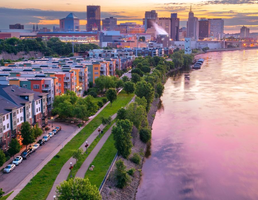 Riverfront cityscape at sunset, colorful sky reflecting on water, with apartments and green trees along a path.